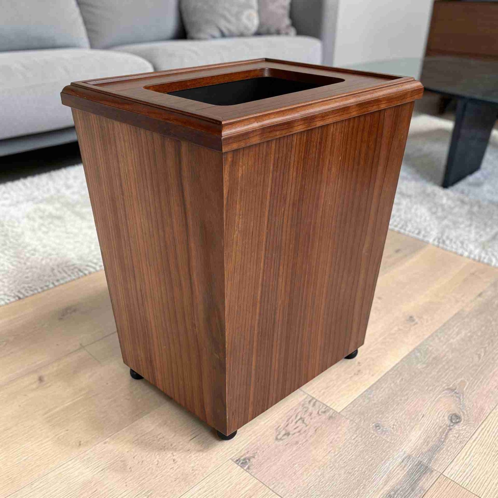 A modern wooden walnut trash can photographed from a slightly low three-quarter angle, placed on a clean, light concrete floor against a subtly textured, off-white studio backdrop with soft directional lighting casting gentle shadows.